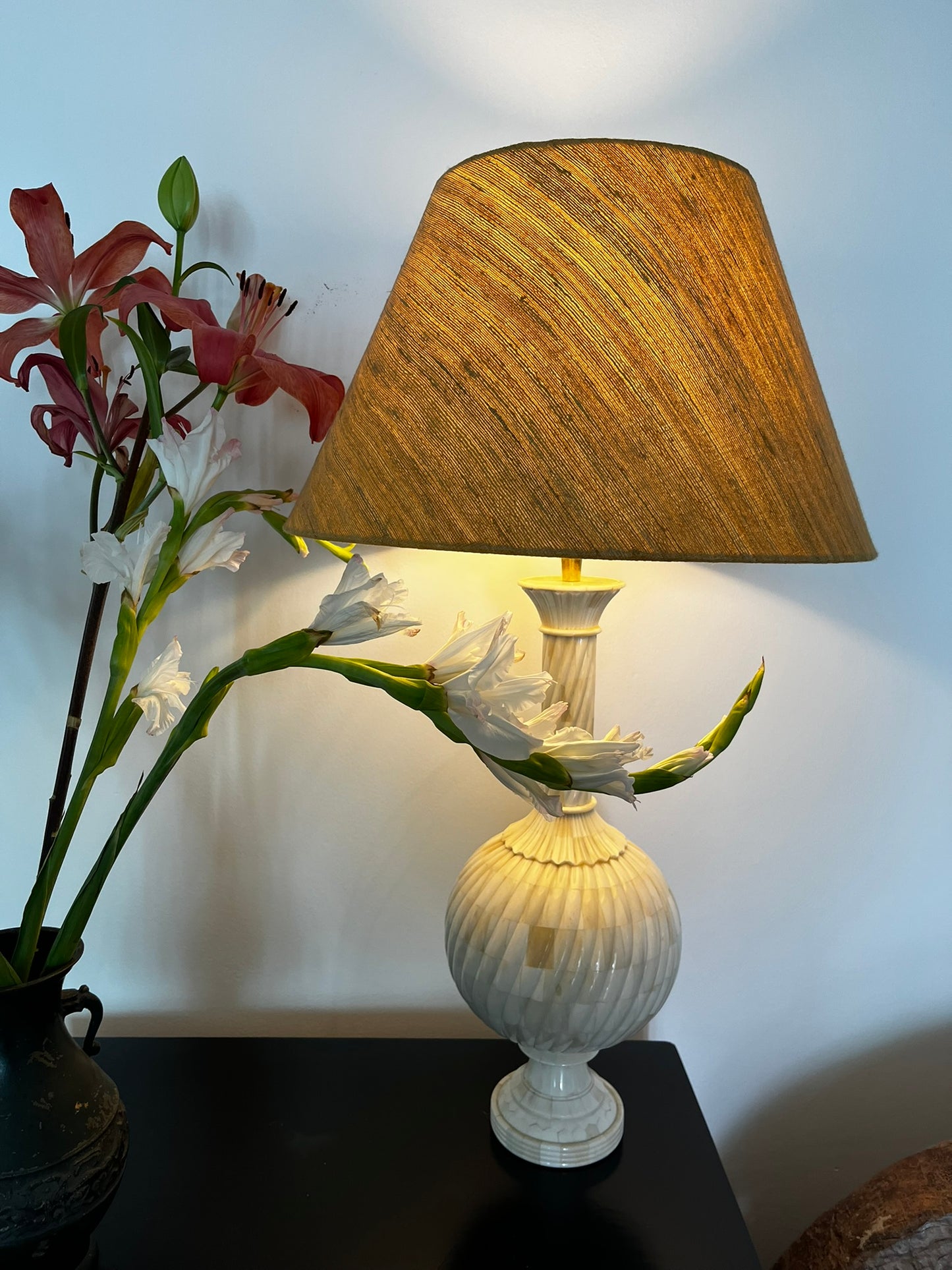 A carved wooden lamp base with bone inlay, intended for use with a lampshade, displayed next to a floral arrangement.