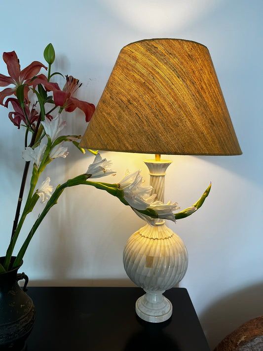 A carved wooden lamp base with bone inlay, intended for use with a lampshade, displayed next to a floral arrangement.