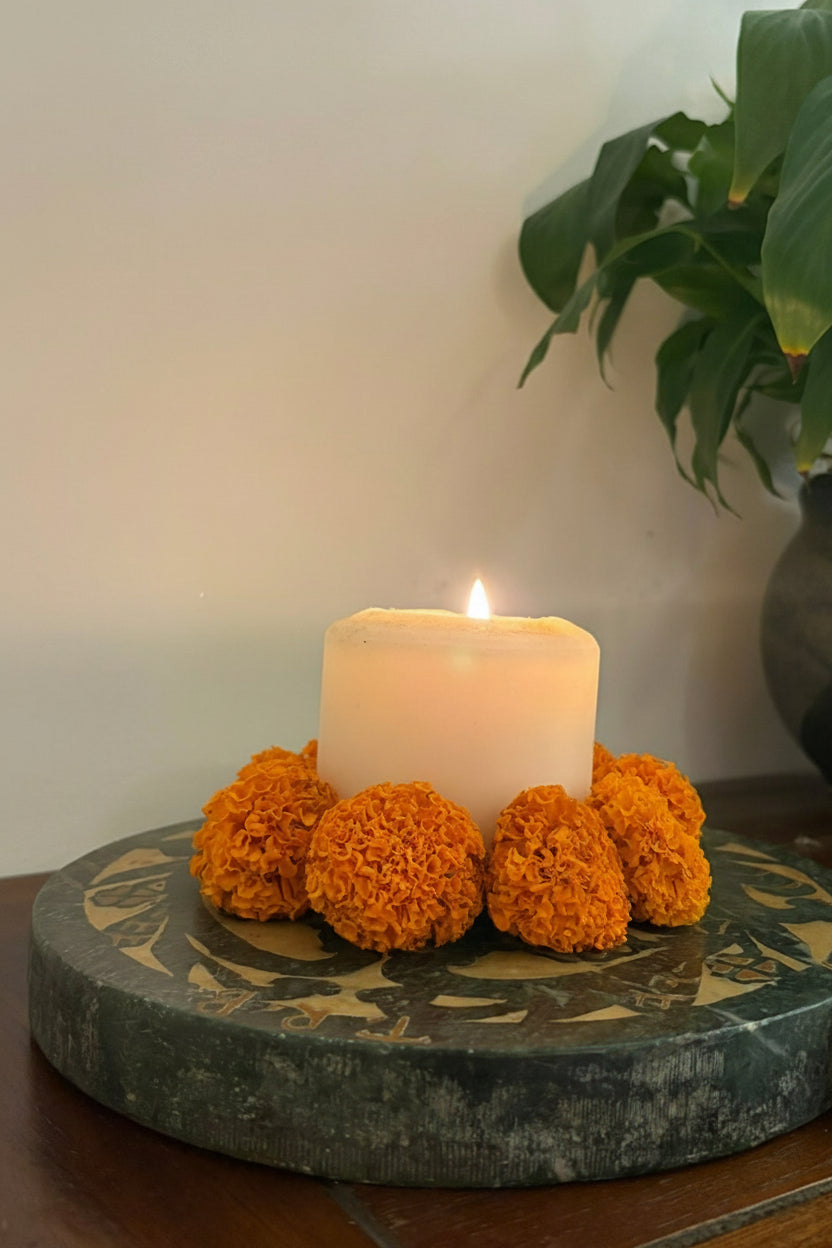 Candle in a decorative holder with orange marigold flowers on a stone vintage candle holder, with a plant in the background.
