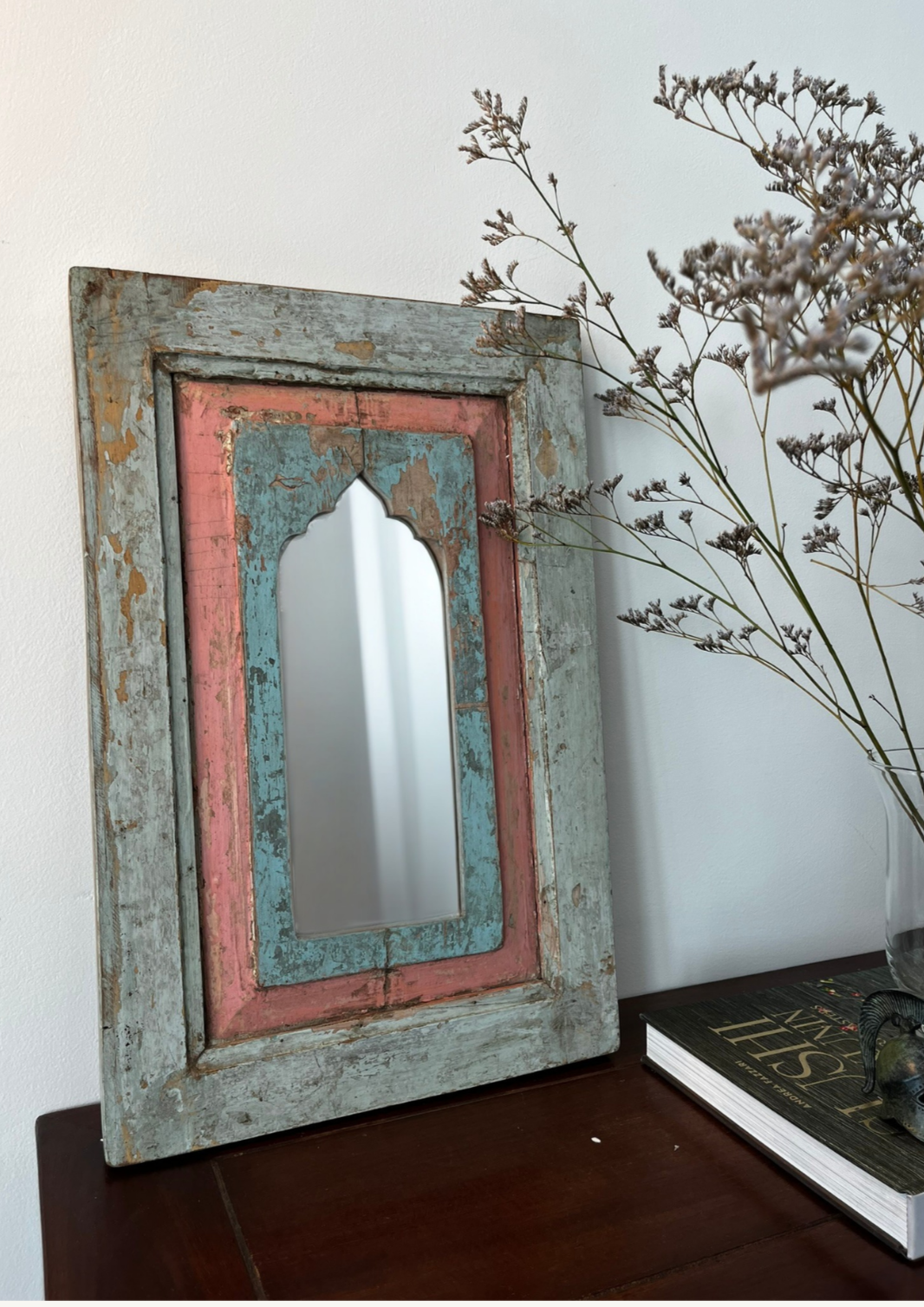 A vintage teakwood mirror with blue and pink accents in an antique style frame, hanging on a wall above a dark wooden surface, with a dried plant to the side.