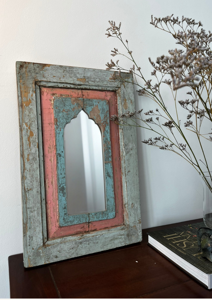 A vintage teakwood mirror with blue and pink accents in an antique style frame, hanging on a wall above a dark wooden surface, with a dried plant to the side.