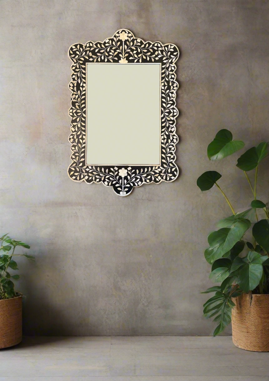 A scalloped, black and white decorative mirror with intricate inlay detailing, hanging on a grey wall between two potted plants.