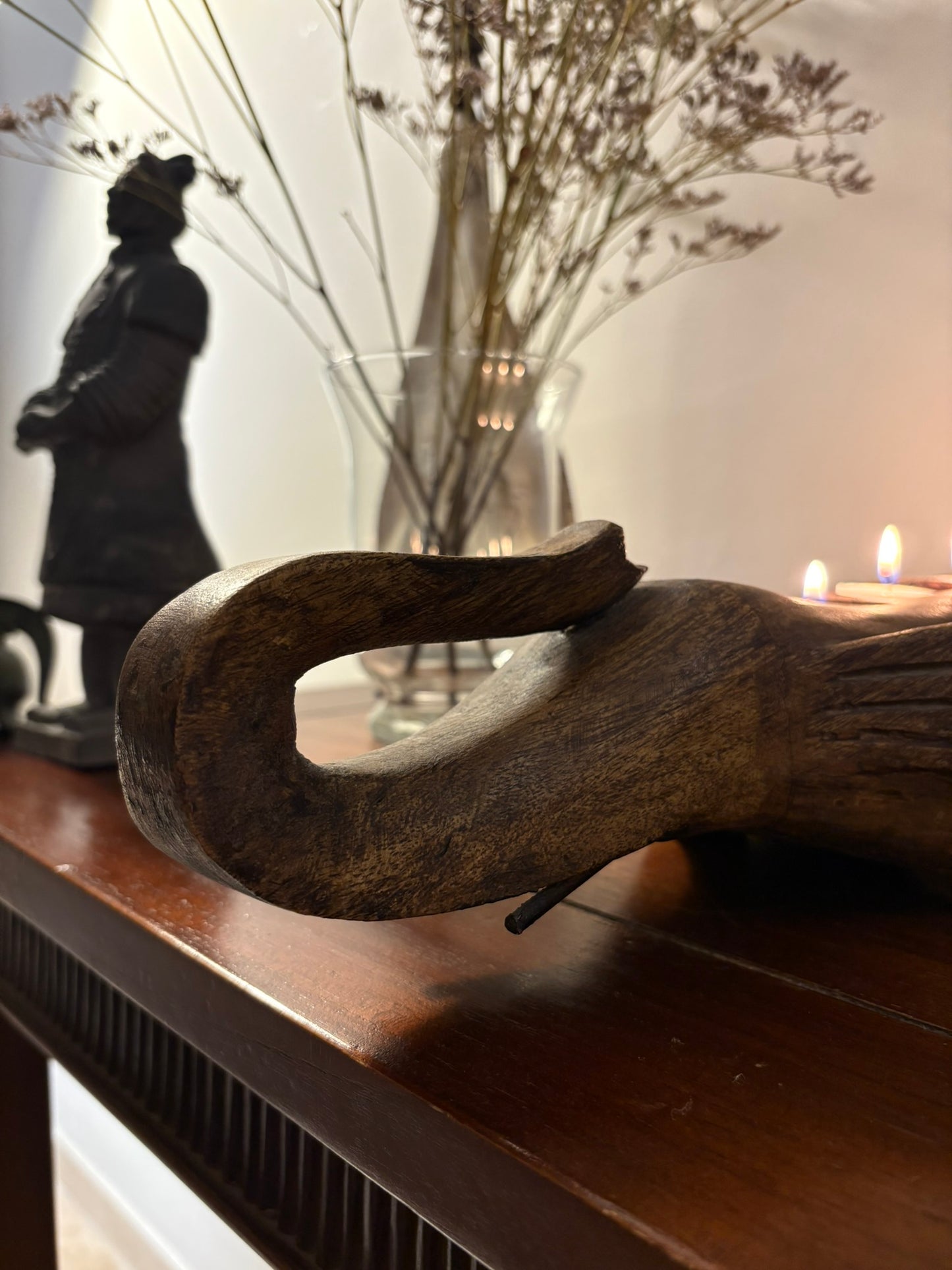 A hand-carved elephant-shaped candle holder made of mango wood, displayed on a table with a silhouette of a person and a vase of dried flowers in the background.