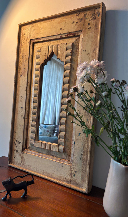 Vintage-style mirror with wooden frame on a wall, next to a vase with flowers and a small figurine.