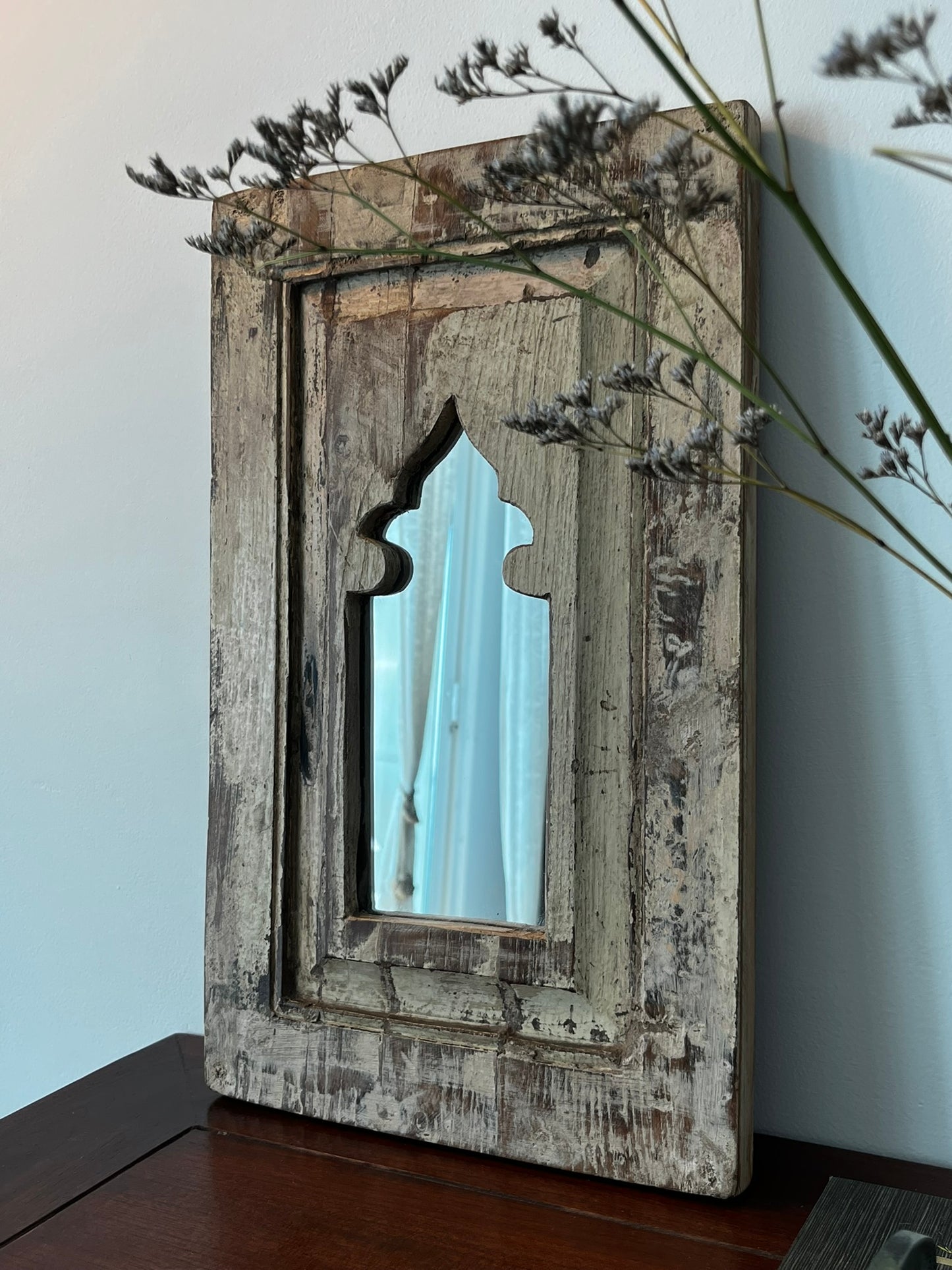 A vintage teakwood mirror with a distinctive aged frame and an arched cut-out in the center, placed against a wall with a plant to the side.