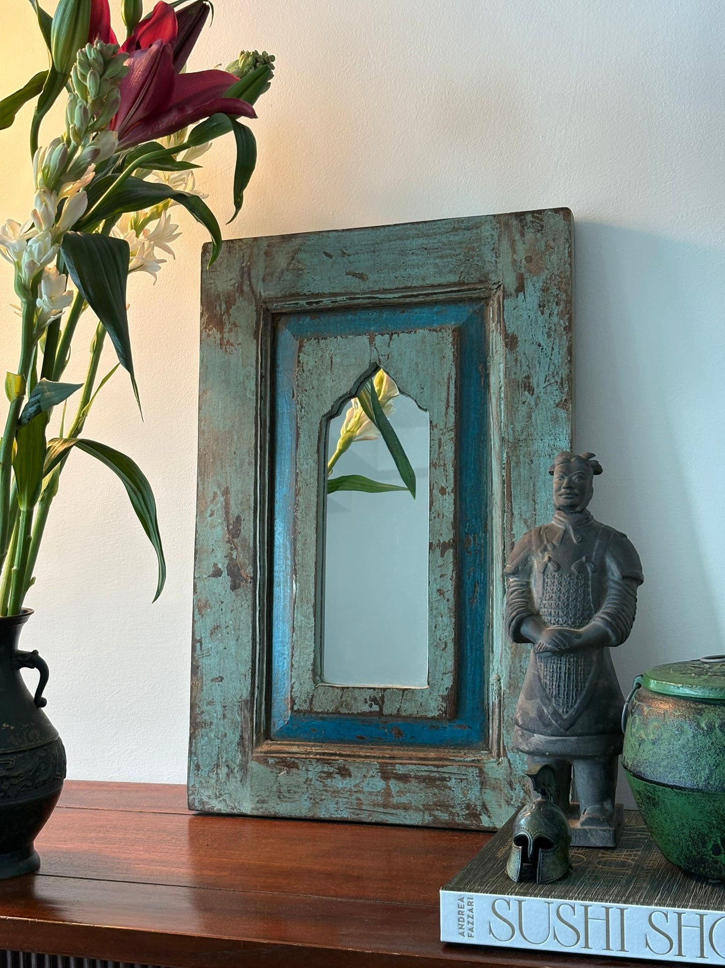 A vintage teakwood mirror with blue trim, hanging on a wall above a table, with a floral arrangement to the left and a statue to the right.