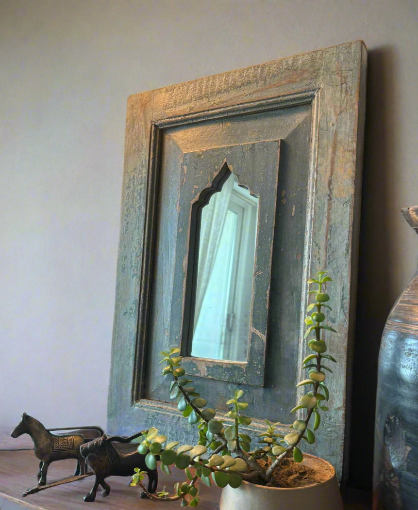 A vintage teakwood mirror with a blue and brown frame, hanging on a wall above a decorative object and a potted plant.
