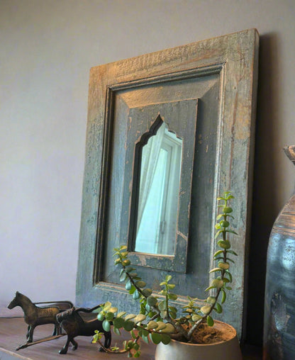A vintage teakwood mirror with a blue and brown frame, hanging on a wall above a decorative object and a potted plant.