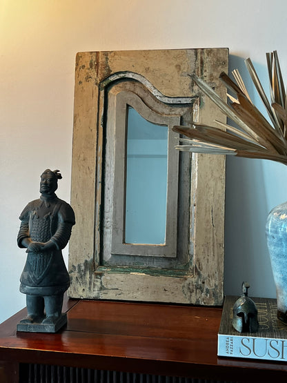 A vintage teakwood mirror with visible signs of aging and use, placed on a wooden surface, with a sculpture and decorative items in the background.